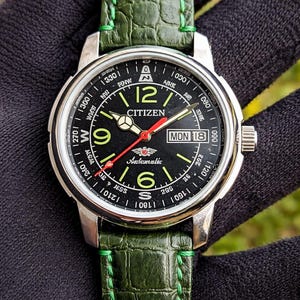 Pode incluir: Um relógio de pulso automático Citizen com mostrador preto, caixa prateada e pulseira de couro verde. O mostrador exibe o dia e a data, com algarismos e ponteiros luminosos. A pulseira verde tem costuras verdes.