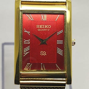 May include: Gold-toned rectangular Seiko Quartz watch with a red face and mesh strap. The watch face features Roman numerals and the Seiko Quartz logo. The watch has a gold-toned case and a mesh strap.