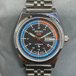 Puede incluir: Un reloj automático Seiko 5 de acero inoxidable con esfera negra, detalles en azul y naranja y visualización de la fecha. El reloj tiene una correa plateada y una esfera redonda. El día es sábado 17.