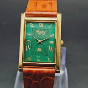 May include: A rectangular gold-toned Seiko Quartz watch with a green face and Roman numeral hour markers. The watch has a brown leather band. The watch face has the Seiko Quartz logo. The watch is a classic timepiece.