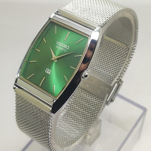 Può includere: Un orologio Seiko Quartz rettangolare con un quadrante verde brillante e lancette e indici argentati. L'orologio presenta un cinturino in maglia d'argento e una cassa argentata. L'orologio è esposto su un supporto trasparente.