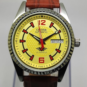 Puede incluir: Un reloj de pulsera automático Citizen vintage con esfera amarilla, detalles rojos y correa de cuero marrón. La esfera muestra el día y la fecha, con el texto "CITIZEN AUTOMATIC" y "21 JEWELS". La caja del reloj es plateada.