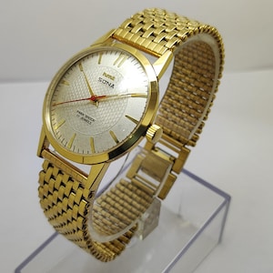 Könnte beinhalten: Goldfarbene Armbanduhr mit strukturiertem silberfarbenem Zifferblatt und goldenem Armband. Das Zifferblatt zeigt den Markennamen "SONA" und den Text "PARA SHOCK 17 JEWELS". Die Uhr hat rote Zeiger und eine goldfarbene Lünette.