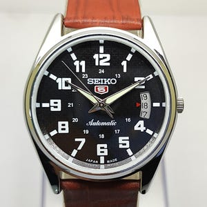 May include: A silver-toned Seiko 5 automatic watch with a black dial, white numerals, and a brown leather strap. The watch face displays the day and date, with the Seiko logo prominently featured. The watch has a stainless steel case.