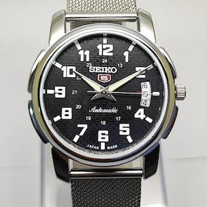 May include: A silver-toned Seiko 5 automatic watch with a black dial. The watch features white numerals, a date display, and a mesh band. The dial includes the text "SEIKO" and "Automatic".