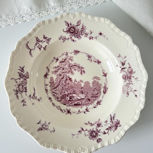 May include: A cream-coloured ceramic bowl with a scalloped edge, featuring a deep purple transferware pattern. The centre depicts a pastoral scene with figures, surrounded by a floral wreath and additional floral sprigs on the rim.
