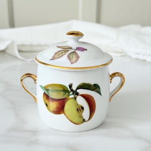May include: White ceramic sugar bowl with lid, featuring a gold rim and handles. The bowl is decorated with a colourful illustration of apples and leaves. The lid has a gold finial and a similar apple and leaf design. A decorative kitchenware item.
