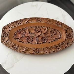 May include: A carved wooden tray with a floral design. The tray is oval-shaped with a border of carved leaves and flowers. The center of the tray features a cluster of carved flowers and leaves. The wood has a warm, brown tone.