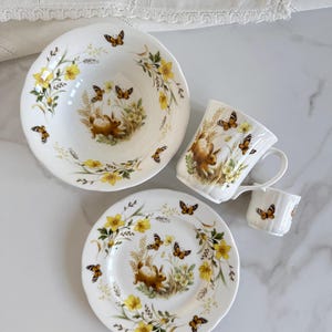 Puede incluir: Juego de vajilla de porcelana blanca con diseño de conejos y mariposas. El juego incluye un cuenco, dos platos, una taza y una taza pequeña. Flores amarillas y follaje verde rodean al conejo y las mariposas.