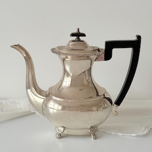 May include: A silver teapot with a black handle and a decorative lid. The teapot has a curved spout and stands on three small feet. The handle is a dark, rectangular shape. The teapot is likely made of metal.