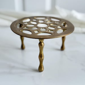 May include: A circular brass trivet featuring an intricate openwork geometric pattern across the top surface. The piece is supported by three slender, curved legs that provide elevation, showcasing a polished metallic finish with visible patina.