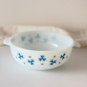 May include: White, round bowl with two handles, decorated with blue and black snowflake designs. The bowl has a smooth, glossy finish and is likely made of ceramic or glass. A vintage kitchenware item.