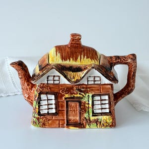 May include: A ceramic teapot shaped like a cottage with a brown thatched roof, a central door, and two windows on the front. The handle is textured like a tree branch, and the spout extends from the left side. The exterior features painted details.