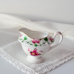 Può includere: Una lattiera in porcellana bianca con un motivo floreale dipinto a mano di rose rosa e foglie verdi. Il recipiente presenta un bordo smerlato con finiture dorate, un manico curvo e una base a piedistallo con dettagli botanici.