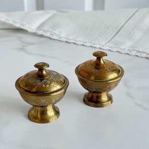 May include: Two small, antique brass bowls with lids. The bowls have a pedestal base and are adorned with etched floral patterns. The lids are topped with a small knob. The bowls are displayed on a marble surface.