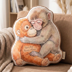 Puede incluir: Un cojín con forma de dos monos abrazados. El mono naranja tiene el texto "You Are Never Alone" impreso. El otro mono es gris y marrón. El cojín tiene un borde marrón.