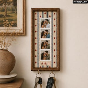 May include: A rectangular wooden wall-mounted key holder featuring a vertical fabric panel with a red apple and blue stripe pattern. The centre displays four stacked photographs of a couple, with the red text A & J below. Two metal hooks hold keys.
