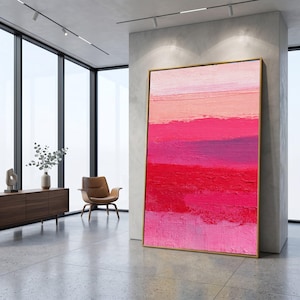 Puede incluir: Gran pintura abstracta con marco dorado. La obra presenta bandas horizontales de colores rosas y rojos. La pintura se apoya en una pared blanca en un espacio interior moderno.