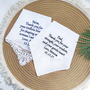 Pode incluir: Dois lenços brancos com mensagens bordadas em azul marinho. Um diz "Mãe, Obrigado pelo seu amor infinito..." e o outro diz "Pai, Obrigado pela sua força..." O lenço da esquerda tem uma borda de renda.