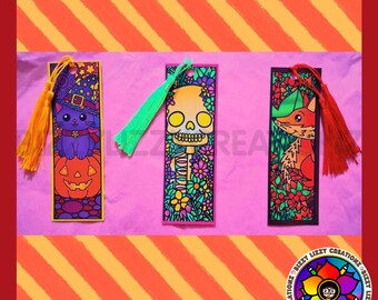 Puede incluir: Tres marcapáginas rectangulares con borlas. El de la izquierda muestra un gato morado con sombrero de bruja sobre una calabaza con una borla naranja. El central presenta una calavera rodeada de flores con una borla verde. El de la derecha muestra un zorro entre follaje con una borla roja.