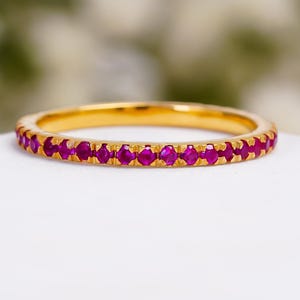 Op de afbeelding: Een dunne gouden ring met een doorlopende rij kleine, ronde, felroze edelstenen die langs de buitenomtrek zijn gezet. Het metaal heeft een gepolijste geelgouden afwerking en de stenen zijn uniform in grootte en kleur over de hele ring.