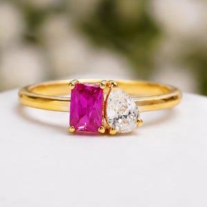 May include: A gold ring featuring two stones set side by side: a vibrant rectangular emerald-cut pink gemstone and a clear pear-shaped gemstone. The band is thin and polished gold, holding the two distinct stones in a prong setting design.