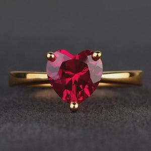May include: A gold ring featuring a faceted heart-shaped red gemstone set in a three-prong mounting. The band is smooth and polished, showcasing a classic design with a vibrant, deep red stone that serves as the central focal point of the piece.