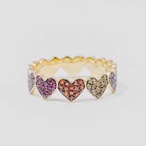 Può includere: Un anello in oro composto da una serie di castoni a forma di cuore collegati tra loro. Ogni cuore è incastonato con piccole pietre preziose dai colori vivaci, in sfumature di rosa, arancione e giallo, creando un motivo a cuore continuo.
