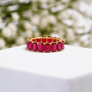 Puede incluir: Un anillo de eternidad con una hilera continua de vibrantes piedras preciosas de corte rectangular en color rojo rosado intenso, engastadas en un metal de tono dorado pulido. El anillo muestra una disposición uniforme de piedras.