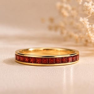 Könnte beinhalten: Ein goldener Bandring mit einer durchgehenden Kanalfassung aus quadratisch geschliffenen, tiefroten Granat-Edelsteinen. Das Metall hat eine polierte Oberfläche und die Steine sind in einer einzigen Reihe um den gesamten Ring angeordnet.
