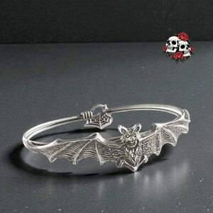 May include: Silver-toned bangle bracelet featuring a detailed bat with outstretched wings. The bat is the focal point, with intricate wing detailing. The bracelet has a spiderweb design near the clasp. A gothic-themed accessory.