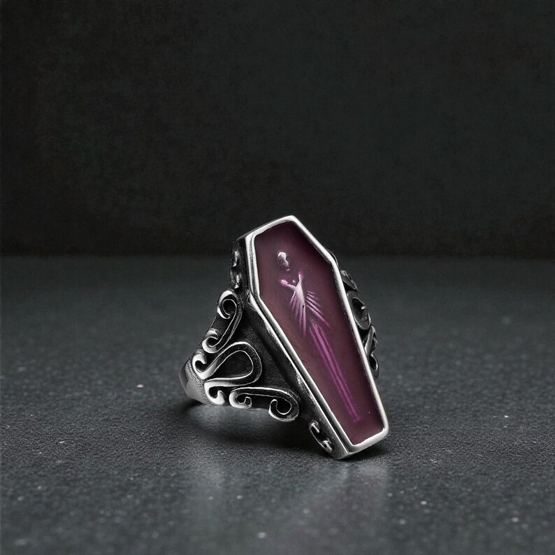 K&ouml;nnte beinhalten: Ein silberner Ring mit Sarg-Design, lila Innenraum und einer Figur. Der Ring hat verzierte Schn&ouml;rkel an den Seiten, was ihm einen gotischen Look verleiht. Der Ring ist vor einem dunklen Hintergrund platziert.