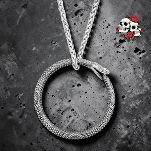 Puede incluir: Un collar de metal plateado con un colgante circular en forma de serpiente que se muerde la cola. La serpiente tiene escamas detalladas y la boca abierta. El colgante cuelga de una cadena de eslabones gruesos de color plateado texturizado.
