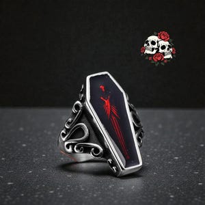 K&ouml;nnte beinhalten: Silberfarbener Ring mit einem Sarg-f&ouml;rmigen Mittelst&uuml;ck mit rotem Design auf schwarzem Hintergrund. Der Ring hat verzierte Seitendetails. Im Hintergrund befindet sich eine kleine Grafik mit Totenk&ouml;pfen und Rosen.