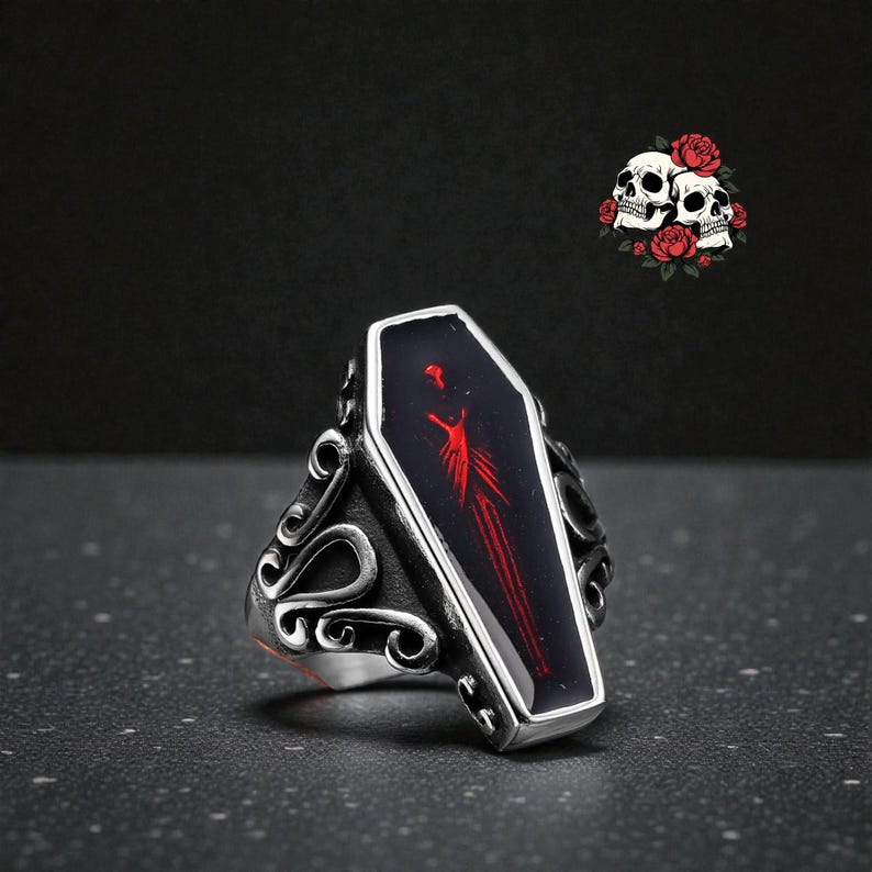 K&ouml;nnte beinhalten: Silberfarbener Ring mit einem Sarg-f&ouml;rmigen Mittelst&uuml;ck mit rotem Design auf schwarzem Hintergrund. Der Ring hat verzierte Seitendetails. Im Hintergrund befindet sich eine kleine Grafik mit Totenk&ouml;pfen und Rosen.