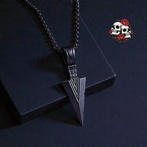 May include: A dark grey arrowhead pendant necklace. The pendant has a triangular shape with etched lines and hangs from a matching chain. The background is a dark blue surface with a small skull and rose graphic.