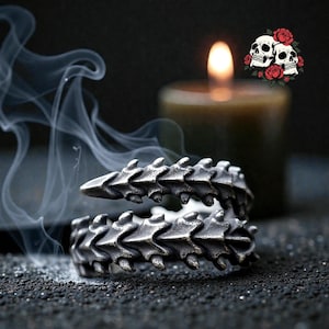 May include: A silver-colored ring with a spine-like design. The ring is wrapped around itself, with the spine segments visible. A lit candle and a skull graphic are in the background, with smoke in the foreground.