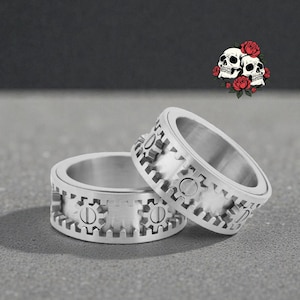 Puede incluir: Dos anillos plateados con diseño de engranaje. Los anillos tienen un acabado de metal cepillado y recortes en forma de engranaje alrededor de la banda. Una pequeña ilustración de calaveras y rosas está en el fondo.