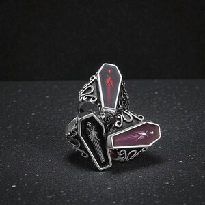 K&ouml;nnte beinhalten: Drei silberfarbene Ringe mit Sarg-f&ouml;rmigen Gesichtern. Jeder Sarg hat eine unterschiedlich farbige Innenseite: schwarz, rot und lila. Die Ringe haben verzierte, wirbelnde Designs um die Sarggesichter, was eine gotische oder alternative &Auml;sthetik erzeugt.