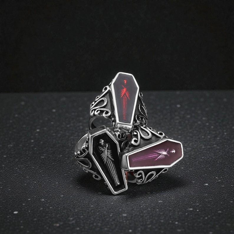 K&ouml;nnte beinhalten: Drei silberfarbene Ringe mit Sarg-f&ouml;rmigen Gesichtern. Jeder Sarg hat eine unterschiedlich farbige Innenseite: schwarz, rot und lila. Die Ringe haben verzierte, wirbelnde Designs um die Sarggesichter, was eine gotische oder alternative &Auml;sthetik erzeugt.