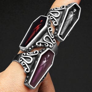 K&ouml;nnte beinhalten: Drei silberfarbene Ringe mit Sarg-f&ouml;rmigen Gesichtern. Ein Ring hat ein rotes Sarggesicht mit rotem Design, ein anderer hat ein schwarz-wei&szlig;es Sarggesicht mit einer Mumie, und der dritte hat ein lila Sarggesicht mit einem Skelett.