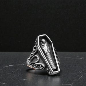 K&ouml;nnte beinhalten: Ein silberfarbener Ring mit einem Sarg-Design und einer Figur darin. Der Ring hat verzierte Schn&ouml;rkel an den Seiten. Der Sarg ist rechteckig mit einer detaillierten Schnitzerei einer verh&uuml;llten Figur.