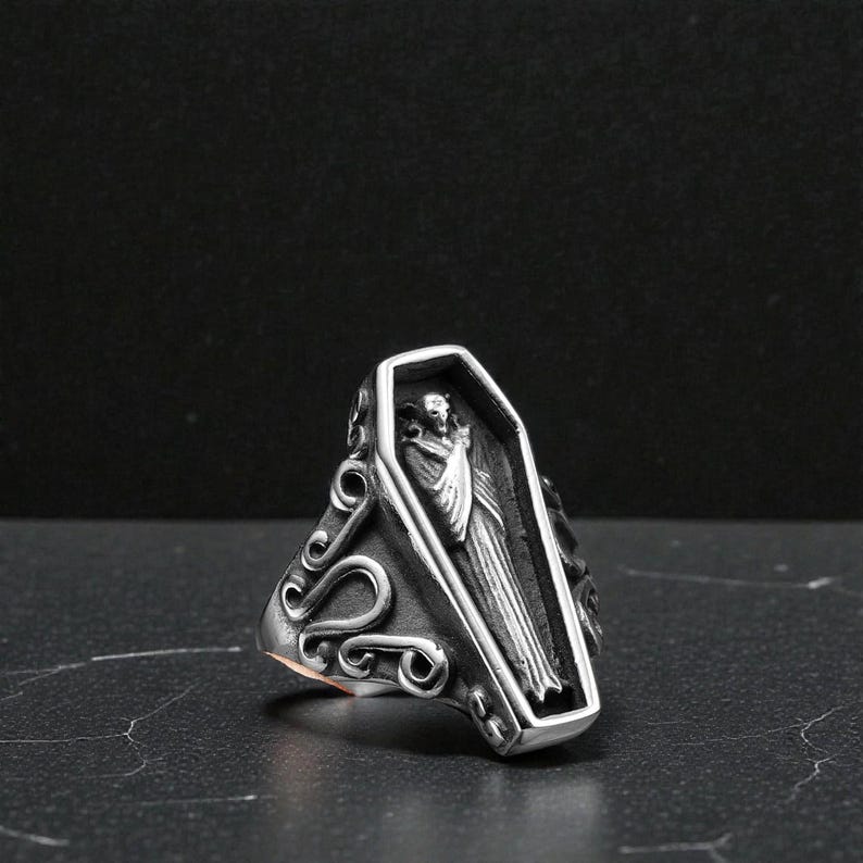 K&ouml;nnte beinhalten: Ein silberfarbener Ring mit einem Sarg-Design und einer Figur darin. Der Ring hat verzierte Schn&ouml;rkel an den Seiten. Der Sarg ist rechteckig mit einer detaillierten Schnitzerei einer verh&uuml;llten Figur.