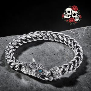 Puede incluir: Una pulsera plateada con un cierre de cabeza de lobo con ojos azules y un diseño de cadena trenzada. La imagen también incluye un gráfico de dos calaveras con rosas rojas.