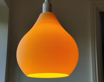 Puede incluir: Una lámpara colgante naranja con una pantalla perforada. La lámpara, con forma de lágrima, emite un brillo amarillo cálido. Cuelga de un cable blanco contra una pared blanca.