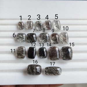 May include: Seventeen polished gemstones are displayed, each with a distinct shape and black inclusions. The stones are numbered 1 to 17, arranged in rows. The gemstones vary in colour, from clear to dark brown.