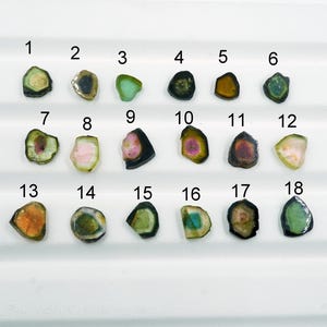 Puede incluir: Una colección de dieciocho rodajas de piedras preciosas pulidas, pequeñas e irregulares, dispuestas en tres filas. Cada piedra está numerada del 1 al 18 y muestra una variedad de colores que incluyen verde, rosa, amarillo y marrón oscuro.