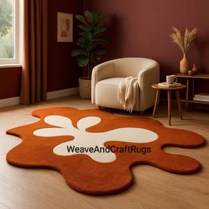 Può includere: Un tappeto moderno dalla forma astratta, con un design color crema su sfondo arancione. Il tappeto è posizionato su un pavimento in legno. Il design del tappeto crea un contrasto sorprendente, aggiungendo un tocco contemporaneo all'arredamento della stanza.