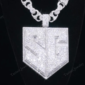 May include: A silver-toned chain necklace featuring a large, shield-shaped pendant covered in clear, sparkling stones. The pendant displays the letters SG in a bold, raised font. The thick chain links are also encrusted with clear stones.