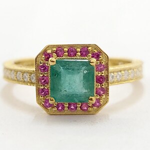 Op de afbeelding: Een gouden ring met een vierkante smaragdgroene middensteen, omringd door kleine roze edelstenen. De ringband is ook van goud, met kleine witte edelstenen langs het oppervlak. Op een witte achtergrond.